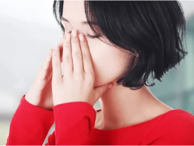 A close-up view of a woman with short dark hair in a red top, holding both hands over her nose and cheeks with her eyes closed, showing facial pain or sinus congestion.