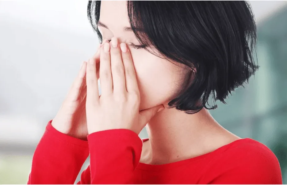 A close-up view of a woman with short dark hair in a red top, holding both hands over her nose and cheeks with her eyes closed, showing facial pain or sinus congestion.